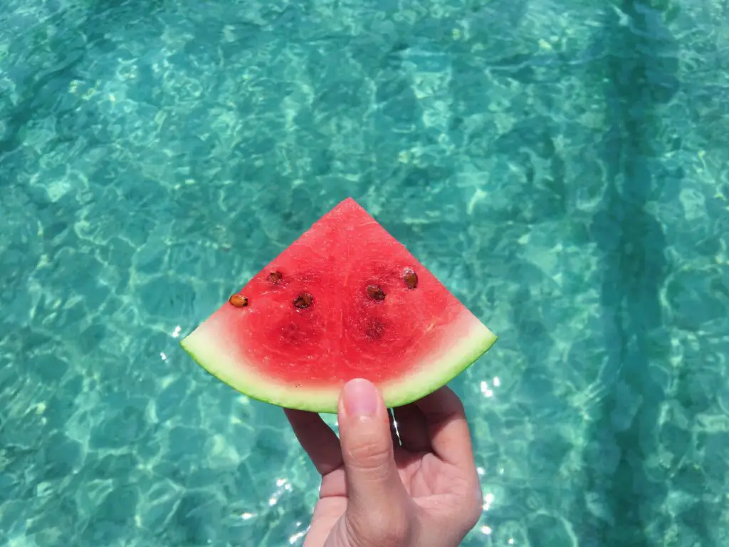 Hand holding a slice of watermelon with blue swimming pool water in the background